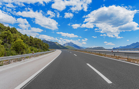 An empty asphalt highway winds through a scenic mountain landscape under a bright blue sky filled with fluffy white clouds.