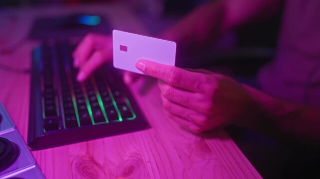 Man holding creditcard with right hand over rgb keyboard while typing in studio; online purchase focus.