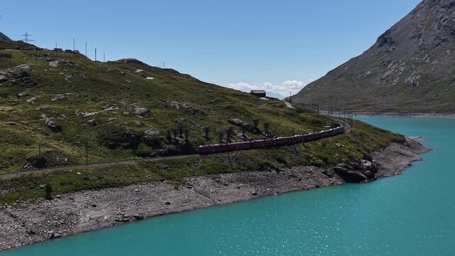 Aerial view of the iconic red Bernina Express train winding along the turquoise waters of Lago Bianco in the mountain landscape of Poschiavo, Grisons, Switzerland.
