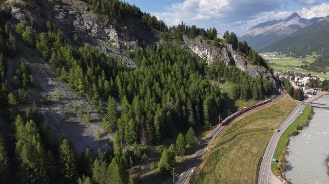 Aerial view of the red Bernina Express train traveling through the mountainous landscape near the river in La Punt Chamues-ch, Grisons, Switzerland.