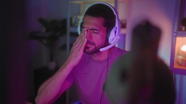 Man wearing white headset rubs eyes with hand while holding gamepad in gaming studio lit by purple led lights; fatigue.