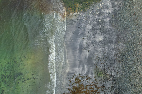 Aerial view of Talisker Bay featuring dark sand, rocky shoreline, and turquoise sea waves crashing against the coast in Talisker, Scotland, United Kingdom.