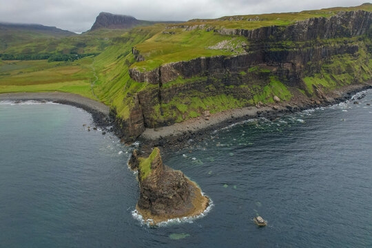 Aerial view of the dramatic sea stack and rugged cliffs of Talisker Bay with a dark pebble beach under a cloudy sky in Talisker, Scotland, United Kingdom.