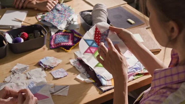 Over the shoulder view of women examining and discussing handmade patchwork quilt blocks at wooden table. Video is suitable for promoting craft workshops, DIY hobby classes, or creative communities