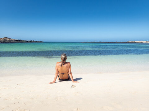 Person Sitting on Sandy Beach Facing Turquoise Ocean