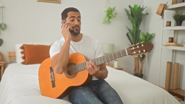 Young hispanic man seated on bed strumming an acoustic guitar and holding smartphone to ear in bedroom with plants and shelves visible; calm focus.