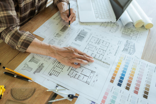 Architect drawing and reviewing detailed building blueprints on a desk with a laptop, rolled plans and color samples, representing architectural design and construction planning.