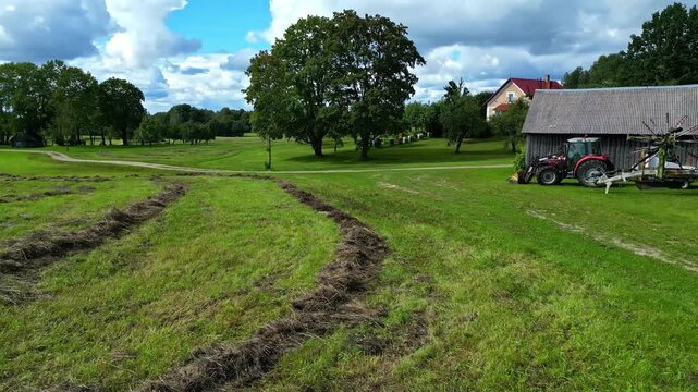 Aerial view of a rural landscape featuring a tractor parked near a barn, rows of mown hay in a green field, and scattered trees under a cloudy sky in Latvia.