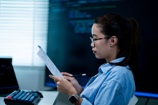 Asian Female Developer with Glasses Reading Printed Document in Dark Office with Code on Screen. Software Engineer Reviewing Technical Documentation.