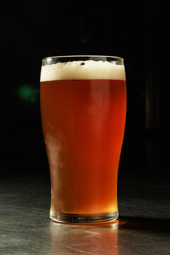 Close-up of a pint glass filled with amber craft beer and a creamy white foam head on a dark bar counter with dramatic studio lighting and a black background. Ideal for brewery, pub, beverage