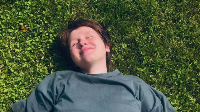 Young person lying on lush green grass in bright sunlight, eyes closed and smiling softly, viewed from overhead with one arm above head and relaxed pose in summer setting
