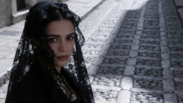 Elegant young woman wearing a traditional black lace mantilla veil outdoors on a cobblestone street, looking at the camera.