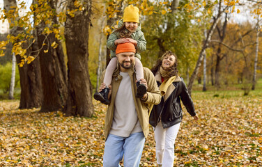 Obraz premium Autumn couple play with leaves in park. Happy parents carry their child on shoulders, enjoying nature and golden foliage during a relaxed stroll. Joyful fall family bonding concept in nature.