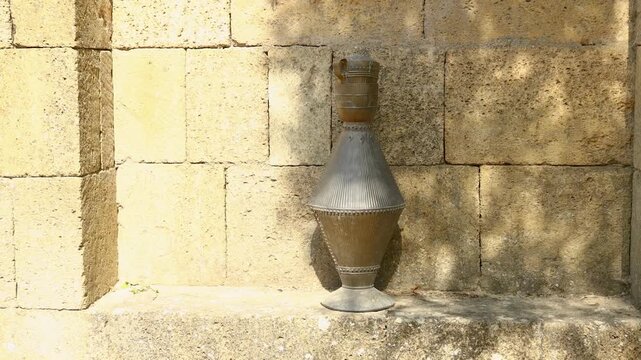 A metal vase sits on a ledge in front of a stone wall, offering a simple yet elegant display