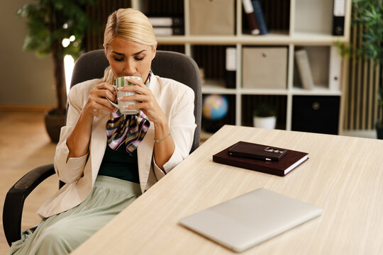 Woman Sits at a Small Office Desk Drinking From a Cup in the Afternoon