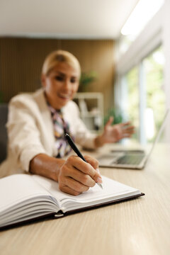 Woman Working in a Small Office Writing Notes While Sitting at a Desk With a Laptop in Front of Her During the Day