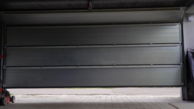 Modern automatic garage door slowly lifting upwards revealing residential suburban street with red brick fence and green grass during day.
