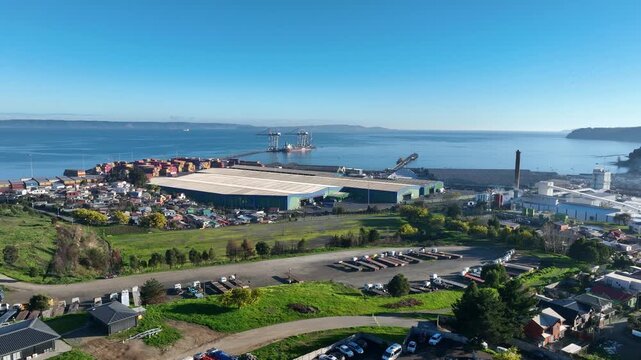 Lateral drift over a coastal industrial town with warehouses, trucks, and parked vehicles overlooking a calm bay. Lirquen, Penco, Chile