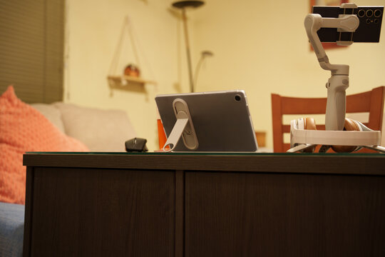 Warm home workspace tabletop with tablet on stand beside smartphone gimbal, soft lamp glow, wooden surface, cozy sofa backdrop, modern remote working setup, with tablet, stand, gimbal, smartphone