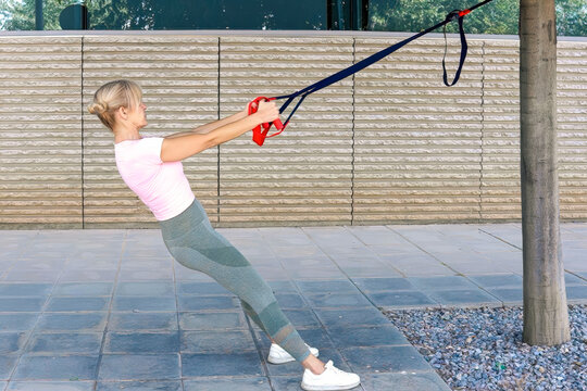 Adult woman performing suspension row workout outdoors with resistance straps anchored overhead, leaning back with straight body, engaged core, flexed arms, focused expression
