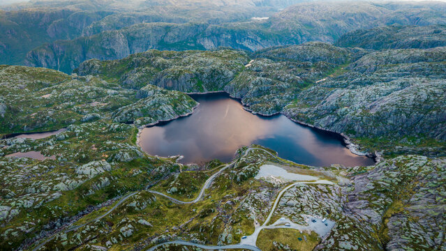 Aerial drone view of a dark mountain tarn and the twisting Lysevegen asphalt road traversing the harsh Norwegian highlands