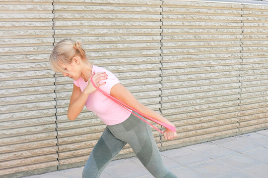Mature woman performing resistance band chest press outdoors beside textured concrete wall in daylight, stepping forward for balance while training upper body strength