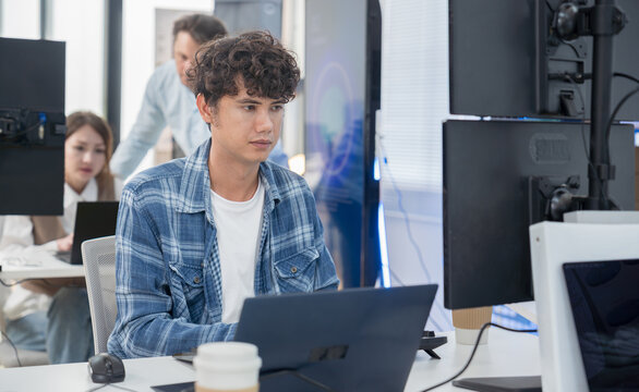 Focused young Asian man uses a laptop at his desk in a high-tech office. A background screen displays an AI assistant interface, representing modern smart technology and digital transformation