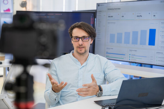 Professional businessman records a financial podcast with a camera, using hand gestures to explain market charts and data analysis in a bright modern studio for investment education.