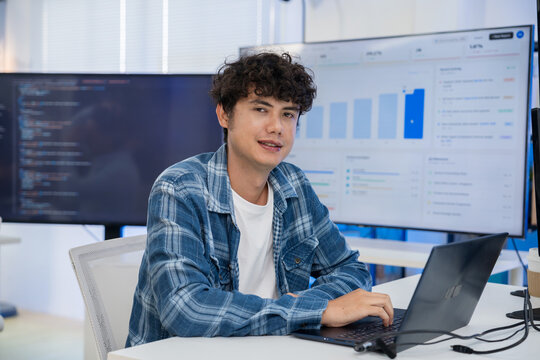 Young Asian technology developer sits at an office desk with a laptop, confidently working near a data analytics screen in a bright modern workplace that reflects innovation and success.