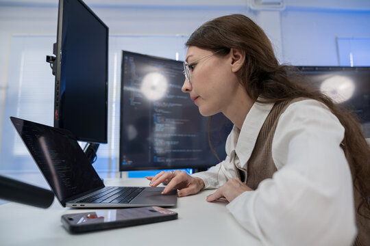 Woman in glasses working on laptop with software code and using smartphone AI voice assistant
