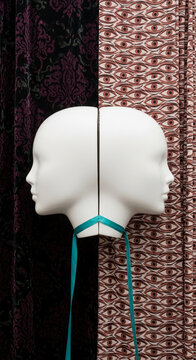 White double-head mannequin with teal ribbon, set against dark floral and eye-patterned fabric, representing duality and perspective.