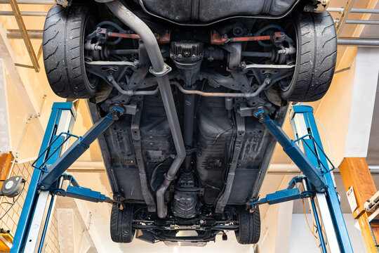 Wide view from directly below showing the complete chassis, exhaust system, and two-post hydraulic lift supporting a race car.