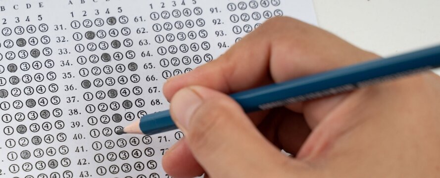 Hand marks answer bubbles on a standardized test sheet. Blue pencil glides over rows of multiple-choice options. Fingers grip the tool with purpose and focus