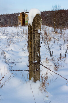 Concrete post wrapped in rusted barbed wire covered with white snow standing in a desolate field with an abandoned building in the background on a cold winter day