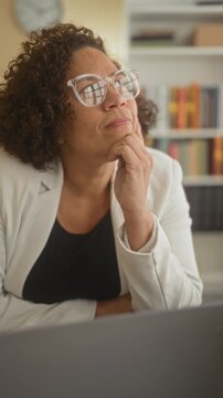 Woman resting chin on hand, wearing glasses, looking away over laptop in a home office with bookshelf and clock; thoughtful reflection.