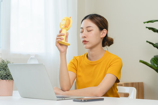 Suffer summer heat wave stroke, hot weather, tired asian young woman sitting on couch in living room without air-conditioner, using electric fan, wave fan to cool for ventilation when high temperature