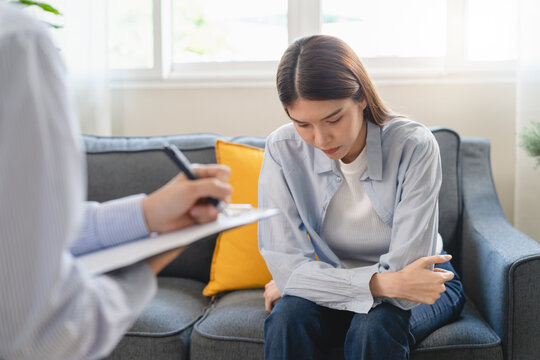 Female psychologist taking notes on clipboard during therapy session with a depressed Asian woman patient