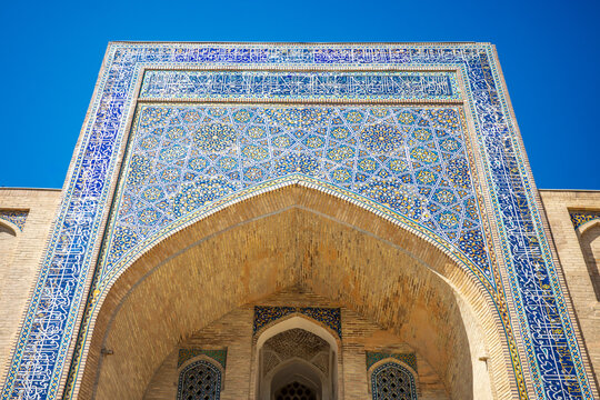 Magnificent historic Islamic entrance in Uzbekistan featuring a monumental pointed arch decorated with intricate blue, turquoise, and gold mosaic tilework, geometric motifs, floral patterns