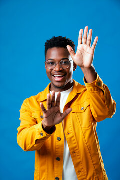 Portrait of young African American man raising hands toward camera and smiling, signaling greeting or stop gesture. Useful for communication, consent, safety, or social campaign themes, vertical