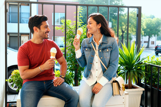 Young adult couple eating ice cream, smiling and chatting during casual date in urban setting, expressing leisure and connection. Ideal for dating, lifestyle, weekend outing marketing