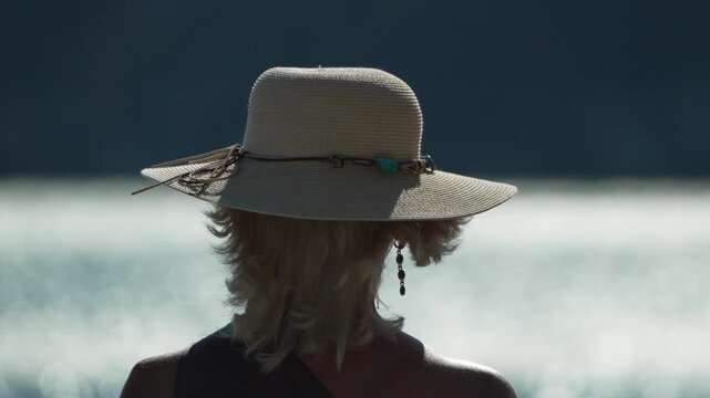 Sun hat woman relaxes by the sea while wind rustles her hair. Medium static camera shot from behind.