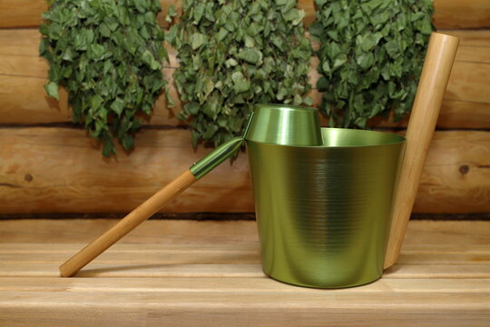 A metal bucket and a ladle are placed on a wooden bench against the backdrop of a log wall, on which dry birch brooms are hanging. 