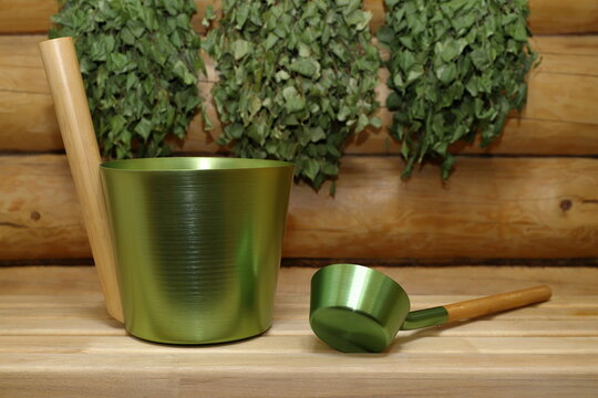 A metal bucket and a ladle are placed on a wooden bench against the backdrop of a log wall, on which dry birch brooms are hanging. 