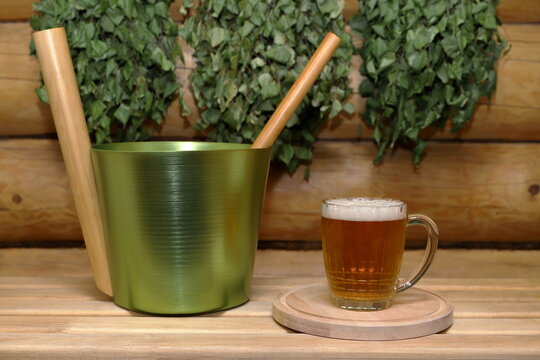On a wooden bench, against the backdrop of a log wall with dry birch brooms hanging on it, there are a mug of light beer on a round wooden tray and a metal bucket with a ladle.