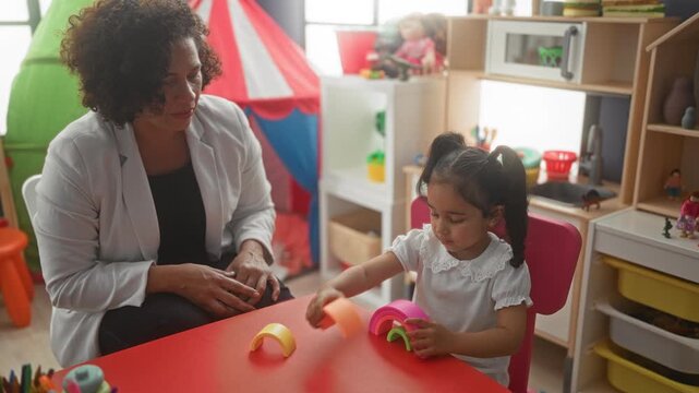 Woman seated beside little girl stacking rainbow toy pieces on a red table in a building playroom filled with tent and shelves; calm learning.