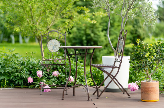Small table and chairs stand on a wooden garden terrace surrounded by lush greenery and potted plants. Cozy outdoor seating area creates a peaceful summer retreat.