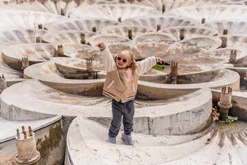 A cheerful little girl is traveling with her family in Armenia, near a sculptural stone fountain with overlapping bowls featuring relief patterns at the entrance to the metro station. © KseniaJoyg