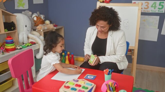 Teacher woman holds letter flashcard as little girl writes with pencil at a red activity table surrounded by toys in a classroom building; concentration.