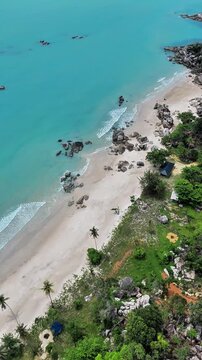 Aerial drone footage of a pristine tropical coastline with crystal turquoise sea, white sandy beach, natural rock formations, palm trees, and lush coastal vegetation creating a peaceful island paradis