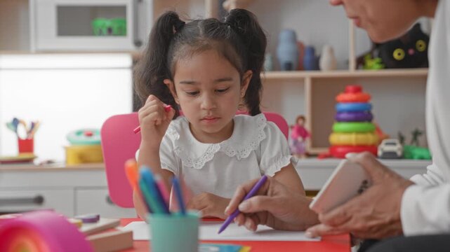 Little girl holding pencil writing at desk in studio while woman teacher gestures and holds a flashcard with number; curiosity learning.